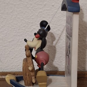 May include: A Mickey Mouse figurine in a boat, with a mast and a sign that says "Mickey Mouse A Steamboat Willie". The figurine is black, red, and yellow. The boat is blue and white.