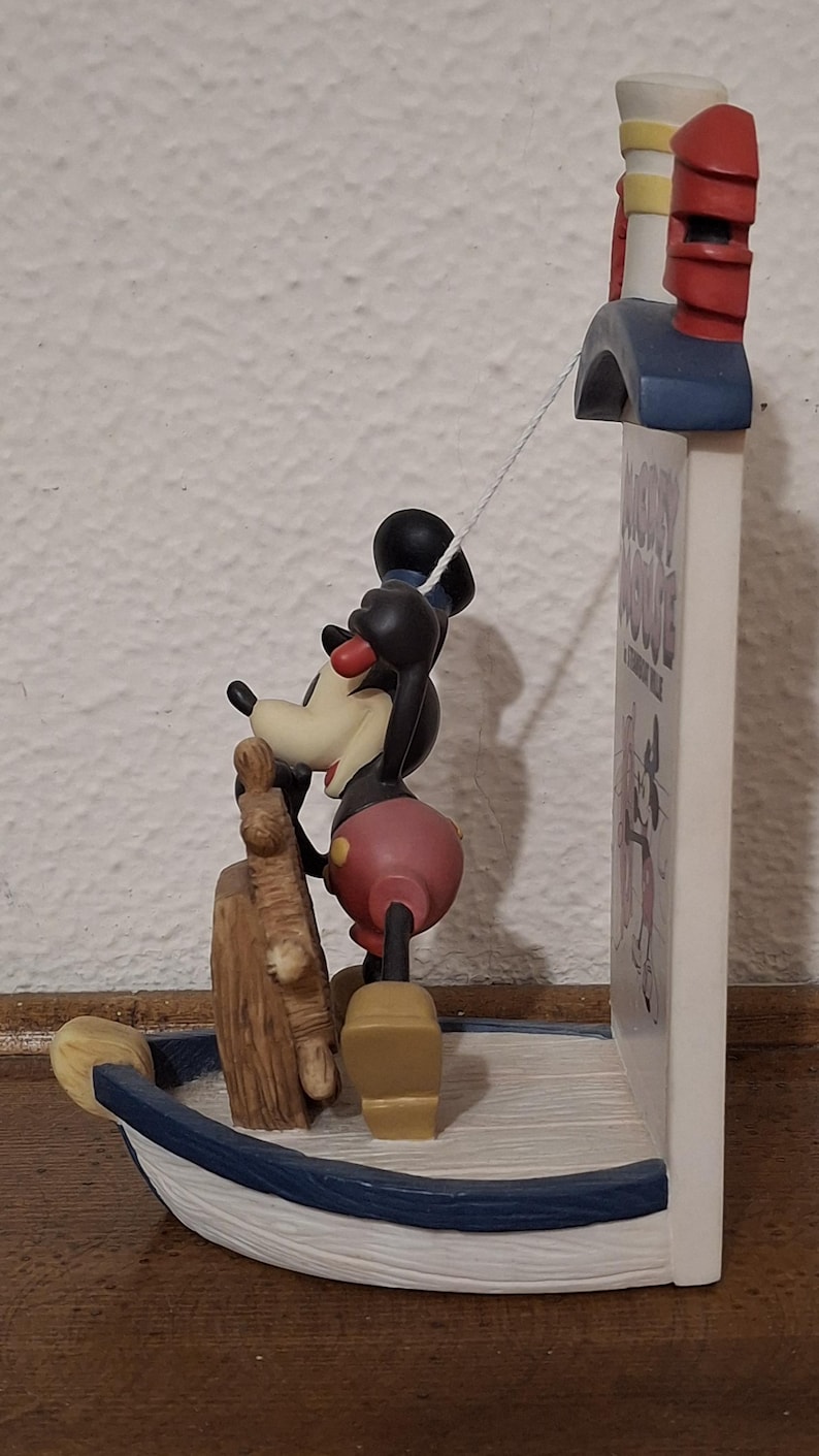 May include: A Mickey Mouse figurine in a boat, with a mast and a sign that says "Mickey Mouse A Steamboat Willie". The figurine is black, red, and yellow. The boat is blue and white.