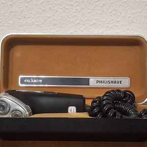 May include: A black electric Philishave shaver with a triple-head rotary blade system and a coiled black power cord, resting inside a rectangular storage case with a tan interior and a silver-trimmed lid featuring the text exclusive PHILISHAVE.
