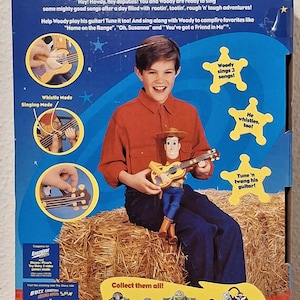 Puede incluir: Caja de la figura de Woody de Toy Story con una guitarra. El embalaje muestra a un ni&ntilde;o sosteniendo la mu&ntilde;eca de Woody, con el texto "Singin' Woody". La caja tambi&eacute;n muestra otros personajes de Toy Story.