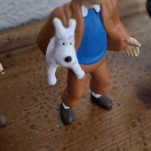 K&ouml;nnte beinhalten: Eine kleine Figur eines hellh&auml;utigen Charakters mit Tolle, bekleidet mit einer braunen Jacke, einem blauen Hemd und einer braunen Hose, der einen kleinen wei&szlig;en Hund mit schwarzen Augen und einer schwarzen Nase h&auml;lt. Die Figur steht aufrecht.