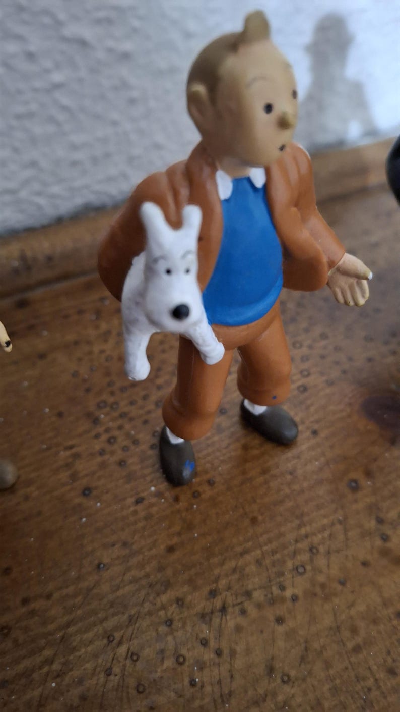 K&ouml;nnte beinhalten: Eine kleine Figur eines hellh&auml;utigen Charakters mit Tolle, bekleidet mit einer braunen Jacke, einem blauen Hemd und einer braunen Hose, der einen kleinen wei&szlig;en Hund mit schwarzen Augen und einer schwarzen Nase h&auml;lt. Die Figur steht aufrecht.