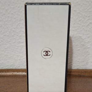 Pode incluir: Caixa de perfume retangular com frente branca e borda marrom. A frente apresenta um logotipo marrom no centro. A caixa est&aacute; sobre uma superf&iacute;cie de madeira.