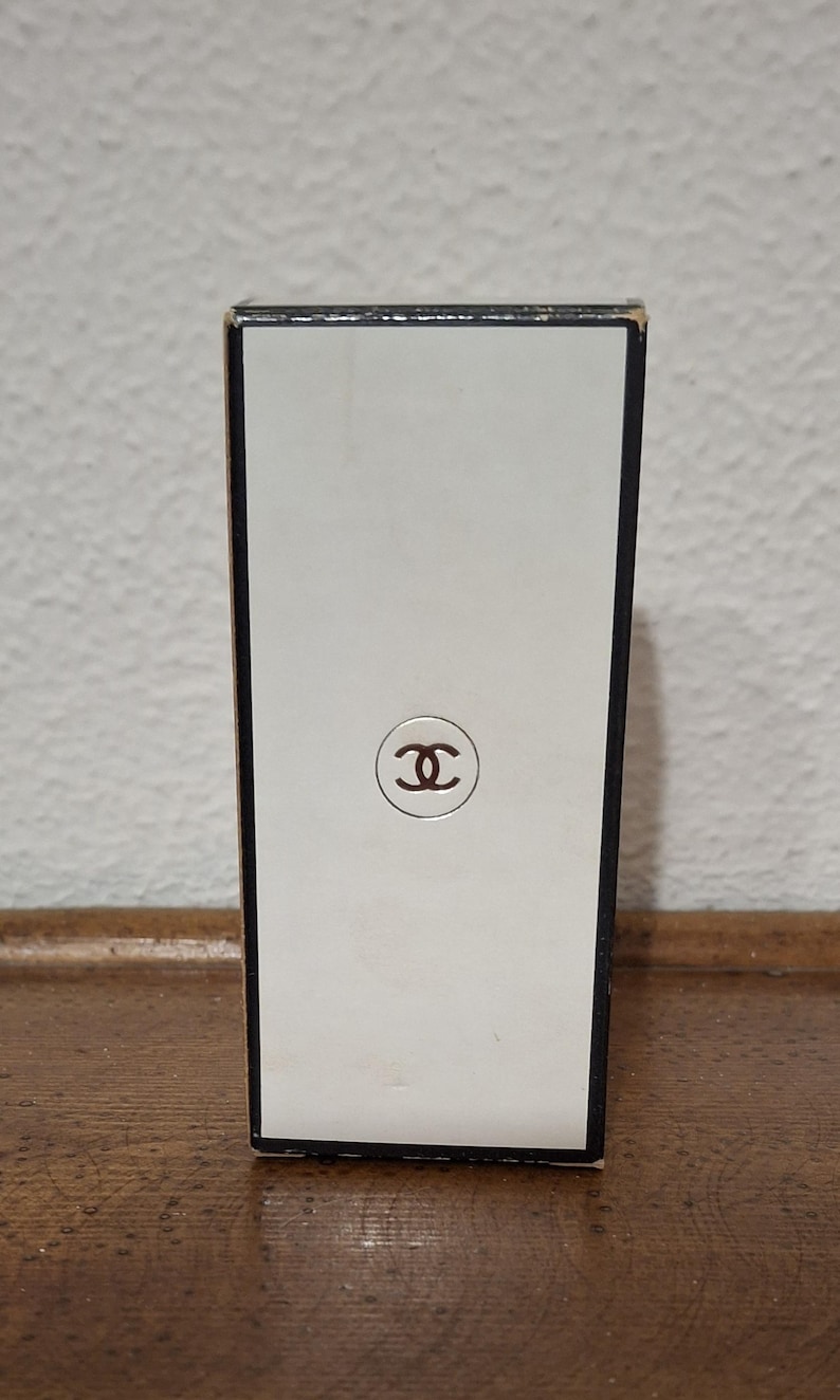 Pode incluir: Caixa de perfume retangular com frente branca e borda marrom. A frente apresenta um logotipo marrom no centro. A caixa est&aacute; sobre uma superf&iacute;cie de madeira.