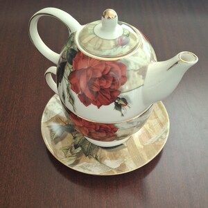 May include: A tea-for-one set consisting of a teapot, a teacup, and a saucer, all featuring a white base with a red rose floral pattern and gold trim. The teapot sits directly on top of the teacup, which rests on the matching saucer.