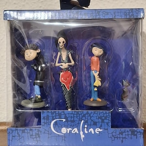 K&ouml;nnte beinhalten: Eine Box mit Coraline-Figuren. Das Set enth&auml;lt drei Figuren: einen Jungen in einem orangefarbenen gestreiften Hemd, eine Frau in einem schwarz-wei&szlig;en Kleid und einen Jungen in einem schwarzen Mantel. Eine schwarze Katzenfigur ist ebenfalls enthalten. Auf der Box steht das Wort "Coraline".