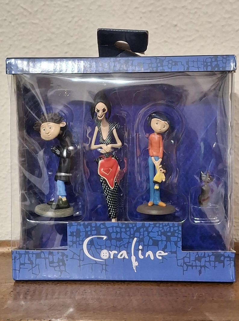 K&ouml;nnte beinhalten: Eine Box mit Coraline-Figuren. Das Set enth&auml;lt drei Figuren: einen Jungen in einem orangefarbenen gestreiften Hemd, eine Frau in einem schwarz-wei&szlig;en Kleid und einen Jungen in einem schwarzen Mantel. Eine schwarze Katzenfigur ist ebenfalls enthalten. Auf der Box steht das Wort "Coraline".