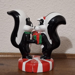 Peut inclure: Figurine en céramique représentant deux moufettes, l'une avec un col vert, l'autre avec un accent rouge, partageant un cadeau. La base est rayée rouge et blanche. Les moufettes sont noires et blanches.