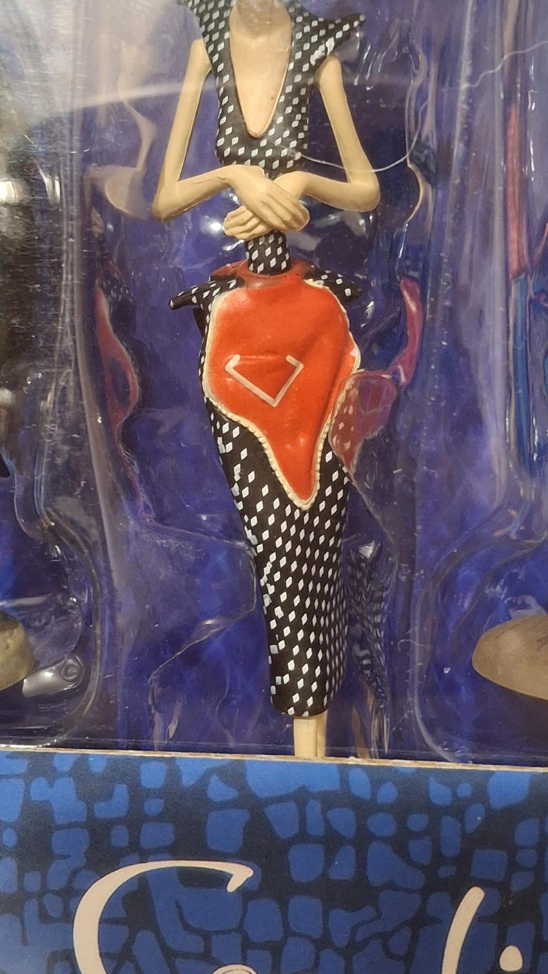 K&ouml;nnte beinhalten: Modepuppe in einer transparenten Kunststoffh&uuml;lle. Die Puppe tr&auml;gt ein schwarz-wei&szlig; gepunktetes Kleid mit einem roten und wei&szlig;en Akzent. Die Arme der Puppe sind verschr&auml;nkt, und sie hat ein einzigartiges, stilisiertes Design.