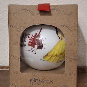 Può includere: Un ornamento sferico bianco con l'illustrazione di un castello rosso e il disegno di un abito giallo, contenuto in una scatola di cartone marrone con apertura circolare e linguetta rossa. La scatola riporta il testo Disney Enchanting Collection.