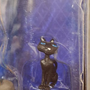 K&ouml;nnte beinhalten: Kleine, schwarze Katzenfigur mit leuchtend blauen Augen und wei&szlig;en Schnurrhaaren. Die Katze sitzt, der Schwanz ist eingerollt. Die Figur befindet sich in einer transparenten Kunststoffverpackung vor blauem Hintergrund.