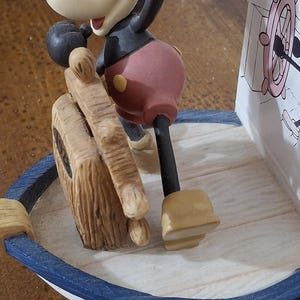May include: A Mickey Mouse figurine in a sailor outfit, standing on a boat-shaped base. The figurine is in a classic pose, holding a wooden ship's wheel. The base has a blue and white trim. A decorative item.