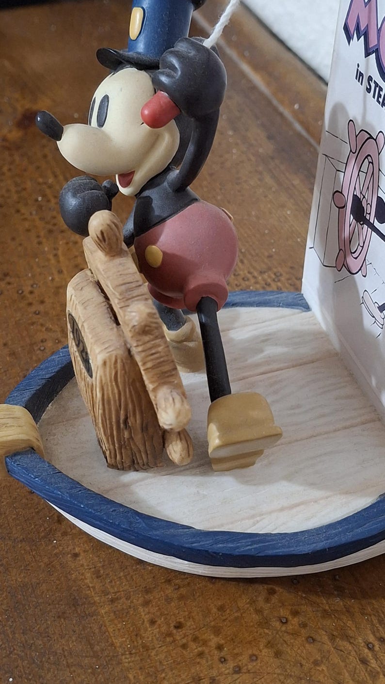 May include: A Mickey Mouse figurine in a sailor outfit, standing on a boat-shaped base. The figurine is in a classic pose, holding a wooden ship's wheel. The base has a blue and white trim. A decorative item.