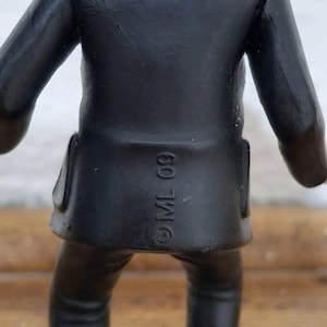 K&ouml;nnte beinhalten: Eine kleine Figur in einem schwarzen Sakko und passender Hose, von hinten betrachtet. Das Sakko hat ein blaues Kragendetail und tr&auml;gt den eingepr&auml;gten Text ML 09 im unteren R&uuml;ckenbereich. Die Figur hat eine glatte, matte Oberfl&auml;che.