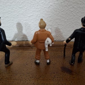 K&ouml;nnte beinhalten: Drei stehende Figuren von hinten betrachtet. Die linke Figur tr&auml;gt eine dunkelblaue Uniform und M&uuml;tze. Die mittlere Figur ist in ein beiges Outfit gekleidet und h&auml;lt ein kleines wei&szlig;es Tier. Die rechte Figur tr&auml;gt einen schwarzen Anzug und Melone.