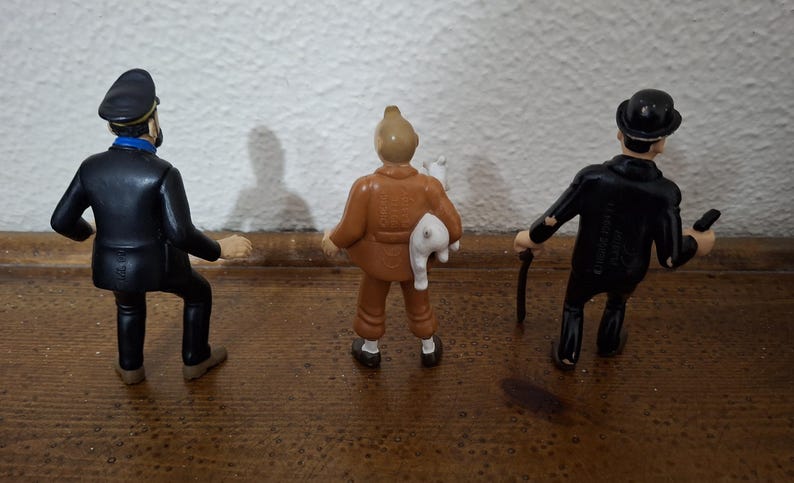 K&ouml;nnte beinhalten: Drei stehende Figuren von hinten betrachtet. Die linke Figur tr&auml;gt eine dunkelblaue Uniform und M&uuml;tze. Die mittlere Figur ist in ein beiges Outfit gekleidet und h&auml;lt ein kleines wei&szlig;es Tier. Die rechte Figur tr&auml;gt einen schwarzen Anzug und Melone.
