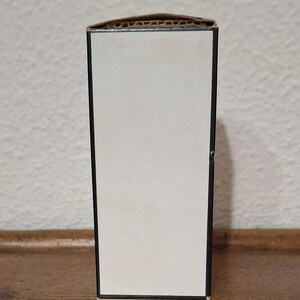 Pode incluir: Uma caixa retangular com frente branca e moldura preta. A borda superior &eacute; feita de papel&atilde;o ondulado. A caixa est&aacute; sobre uma superf&iacute;cie de madeira.