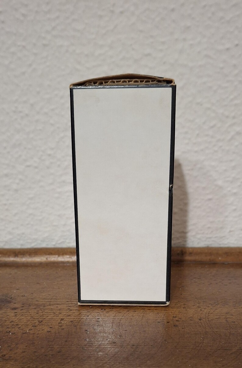 Pode incluir: Uma caixa retangular com frente branca e moldura preta. A borda superior &eacute; feita de papel&atilde;o ondulado. A caixa est&aacute; sobre uma superf&iacute;cie de madeira.