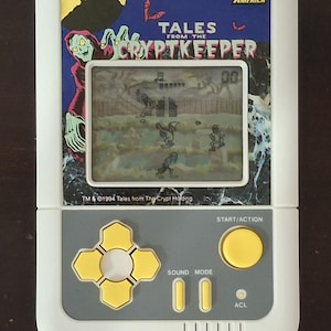 Peut inclure: Jeu électronique portable blanc avec le titre "Tales from the Cryptkeeper". Le jeu a un petit écran affichant une scène pixélisée, avec des boutons jaunes et un pavé directionnel. Le design comprend un personnage de Cryptkeeper et le logo Micro Games of America.