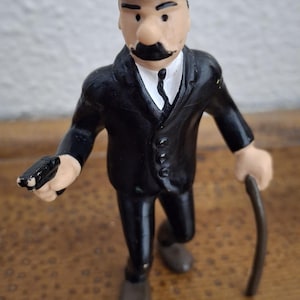 K&ouml;nnte beinhalten: Eine kleine Spielfigur, die eine Person in einem schwarzen Anzug, einem wei&szlig;en Hemd mit schwarzer Krawatte und einer schwarzen Melone darstellt. Die Figur hat einen markanten schwarzen Schnurrbart, h&auml;lt eine kleine schwarze Pistole und einen Gehstock.