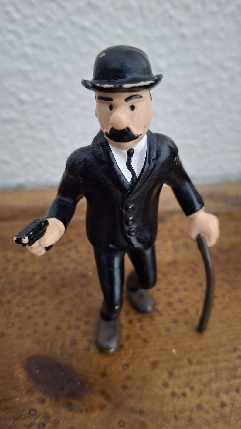 K&ouml;nnte beinhalten: Eine kleine Spielfigur, die eine Person in einem schwarzen Anzug, einem wei&szlig;en Hemd mit schwarzer Krawatte und einer schwarzen Melone darstellt. Die Figur hat einen markanten schwarzen Schnurrbart, h&auml;lt eine kleine schwarze Pistole und einen Gehstock.