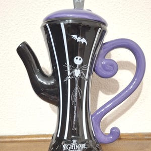 Pode incluir: Bule de cerâmica preto e branco com Jack Skellington de O Estranho Mundo de Jack. O bule tem uma alça e tampa roxas, com uma lápide RIP. O design inclui listras brancas e um morcego.