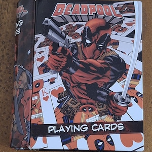 May include: A rectangular Deadpool playing cards deck box featuring a graphic illustration of the character holding a silver handgun and a katana. The background displays multiple playing cards with heart suits. The text DEADPOOL and PLAYING CARDS are printed on the front.