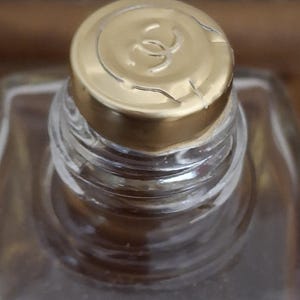 Pode incluir: Close-up de um frasco de perfume com tampa dourada. A tampa apresenta um log&oacute;tipo e est&aacute; ligeiramente rachada. O frasco &eacute; feito de vidro transparente e cont&eacute;m um l&iacute;quido castanho claro. O foco est&aacute; na parte superior do frasco.