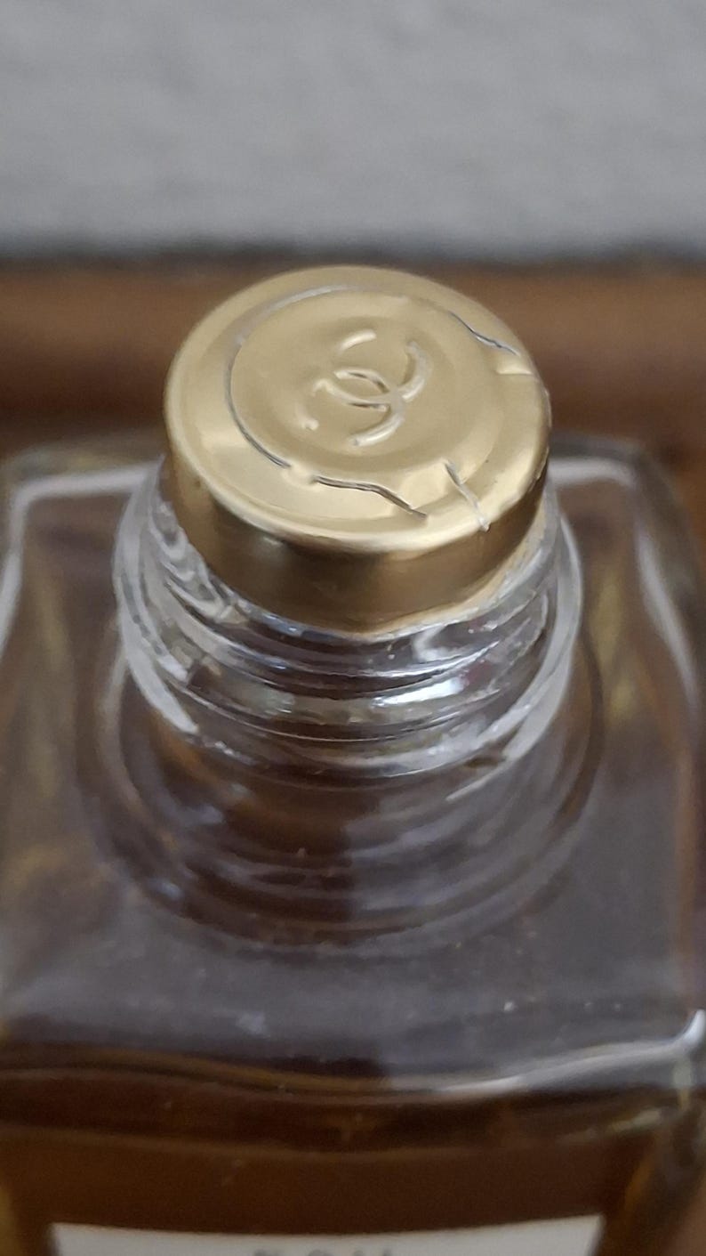 Pode incluir: Close-up de um frasco de perfume com tampa dourada. A tampa apresenta um log&oacute;tipo e est&aacute; ligeiramente rachada. O frasco &eacute; feito de vidro transparente e cont&eacute;m um l&iacute;quido castanho claro. O foco est&aacute; na parte superior do frasco.