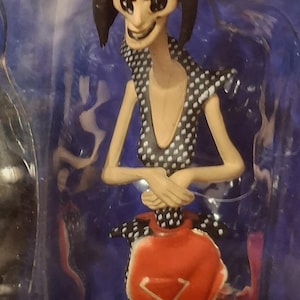 K&ouml;nnte beinhalten: Figur einer Figur mit blasser Haut, schwarzen Haaren und gro&szlig;en schwarzen Augen. Die Figur tr&auml;gt ein schwarzes Kleid mit wei&szlig;en Punkten und ein rotes sch&uuml;rzenartiges Kleidungsst&uuml;ck. Die Arme der Figur sind verschr&auml;nkt, und sie hat ein finsteres L&auml;cheln.