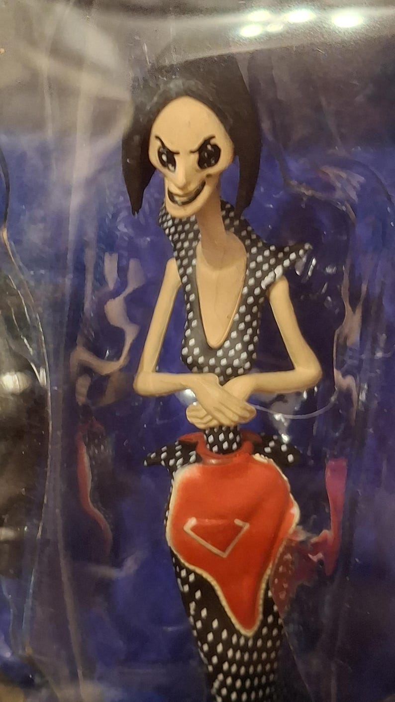 K&ouml;nnte beinhalten: Figur einer Figur mit blasser Haut, schwarzen Haaren und gro&szlig;en schwarzen Augen. Die Figur tr&auml;gt ein schwarzes Kleid mit wei&szlig;en Punkten und ein rotes sch&uuml;rzenartiges Kleidungsst&uuml;ck. Die Arme der Figur sind verschr&auml;nkt, und sie hat ein finsteres L&auml;cheln.