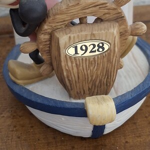 May include: A nautical-themed figurine featuring Mickey Mouse at the helm of a boat. The boat has a blue and white striped design. The wooden helm has the year "1928" on it. The figurine is a collectible item.