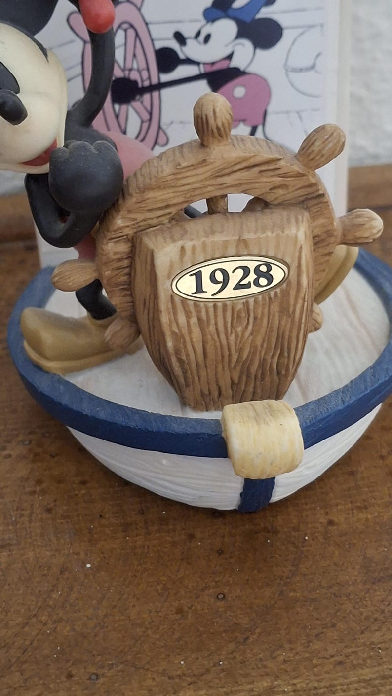 May include: A nautical-themed figurine featuring Mickey Mouse at the helm of a boat. The boat has a blue and white striped design. The wooden helm has the year "1928" on it. The figurine is a collectible item.