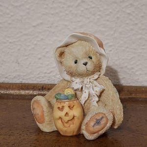 Op de afbeelding: Een beige teddybeerbeeldje met een witte muts en een strik met gouden stippen. De beer houdt een uitgesneden pompoen met een lachend gezicht vast. Het beeldje heeft een vintage, verzamelbaar uiterlijk.