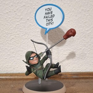 May include: A figurine of a character in a green hooded suit holding a bow with a red boxing glove attached to the string. Above the figure is a blue-bordered speech bubble containing the text YOU HAVE FAILED THIS CITY! The figure stands on a circular base.