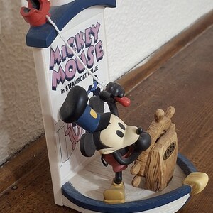 May include: A figurine of Mickey Mouse in Steamboat Willie. The figurine features Mickey Mouse in a sailor outfit, leaning against a wooden ship wheel. The base is blue and white, with the text "Mickey Mouse" on a sign.