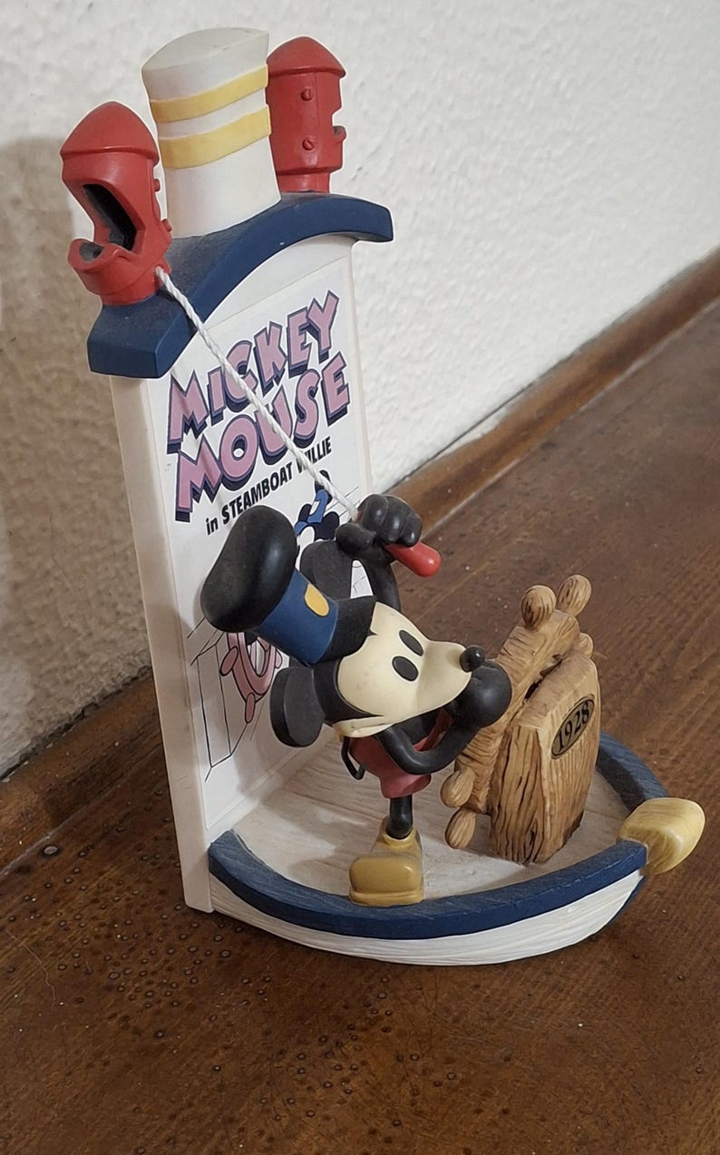 May include: A figurine of Mickey Mouse in Steamboat Willie. The figurine features Mickey Mouse in a sailor outfit, leaning against a wooden ship wheel. The base is blue and white, with the text "Mickey Mouse" on a sign.