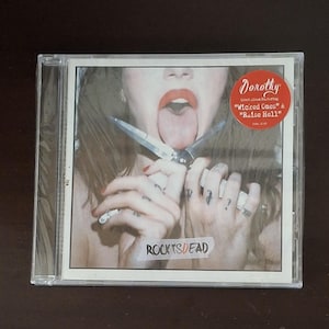 May include: CD case featuring the album "Rocks is Dead" by Dorothy. The cover art shows a woman with her tongue out, holding a knife. A sticker on the case reads "Dorothy" and lists the songs "Wicked Ones" and "Raise Hell."