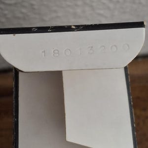 Pode incluir: Close-up de uma caixa de cart&atilde;o branca parcialmente aberta com o n&uacute;mero "18013200" impresso na aba interna. A caixa tem uma borda preta e est&aacute; sobre uma superf&iacute;cie de madeira.