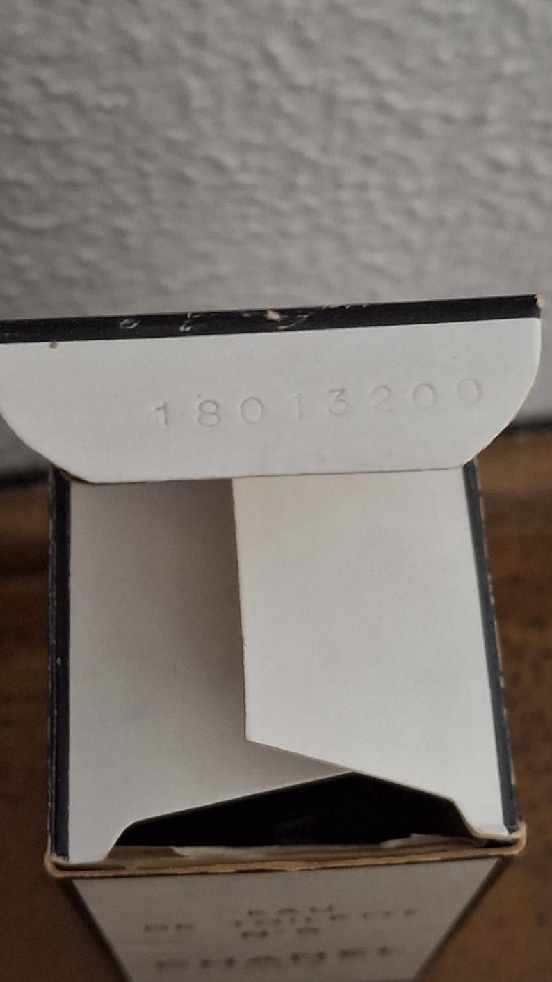 Pode incluir: Close-up de uma caixa de cart&atilde;o branca parcialmente aberta com o n&uacute;mero "18013200" impresso na aba interna. A caixa tem uma borda preta e est&aacute; sobre uma superf&iacute;cie de madeira.