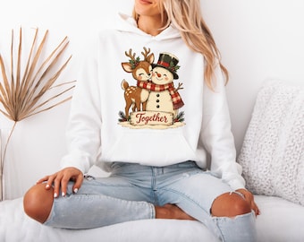 May include: A white hooded sweatshirt featuring a graphic print of a brown deer hugging a snowman wearing a black top hat and a red plaid scarf. The graphic includes the word Together written on a wooden sign at the base of the figures.