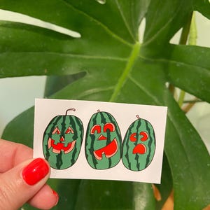 May include: A rectangular sticker sheet featuring three green watermelon illustrations carved like jack-o'-lanterns. Each watermelon has a unique facial expression with bright red cutouts for eyes, noses, and mouths, mimicking the style of Halloween pumpkins.