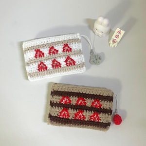 以下が含まれることがあります： 赤い花の模様の、かぎ針編みのポーチが2つ。1つは白地にベージュと赤のアクセント、もう1つは茶色地にベージュと赤のアクセント。各ポーチには小さなループと装飾的なチャームが付いています。小さな白いウサギの置物とイチゴ型のアイテムも存在します。