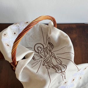 May include: A woven basket with a brown and tan handle holds a cream-coloured cloth. The cloth features an embroidered image of a figure with a halo, along with decorative floral and geometric patterns. The basket and cloth are on a dark wooden surface.