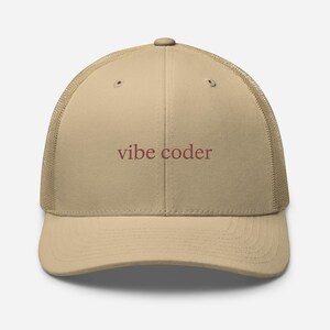 Puede incluir: Gorra de camionero beige con parte trasera de malla y visera curva. La gorra tiene el texto "vibe coder" en burdeos. Un accesorio casual.