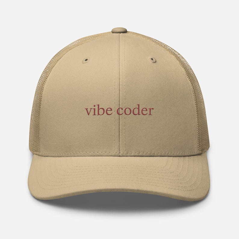 Puede incluir: Gorra de camionero beige con parte trasera de malla y visera curva. La gorra tiene el texto "vibe coder" en burdeos. Un accesorio casual.