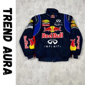 Puede incluir: Chaqueta de carreras Red Bull azul marino con logotipos de patrocinadores, incluyendo Infiniti y Pepe Jeans. La chaqueta tiene cremallera completa, cuello alto y detalles rojos y amarillos. El texto "TREND AURA" está en el lado izquierdo.