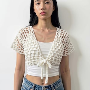 May include: A white short-sleeved crochet bolero with an open-knit net pattern and a front tie closure, worn over a plain white cropped tank top and blue denim jeans. The bolero features a V-neckline and textured yarn construction.