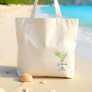 May include: Cream-colored tote bag with a margarita graphic. The margarita is green and has a lime wedge. The name "Hannah" is written in cursive below the margarita. The bag has two handles and is sitting on a sandy beach.