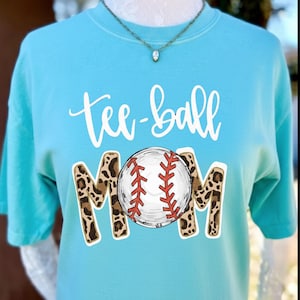 May include: A light blue t-shirt with the words "tee-ball MOM" in white script and leopard print lettering. A baseball graphic is centered on the shirt, featuring red stitching.